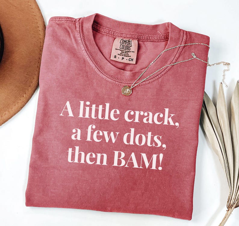May include: A red-toned t-shirt with the text "A little crack, a few dots, then BAM!" in white. A silver necklace with a small pendant rests on the shirt. The shirt has a Comfort Colors tag.
