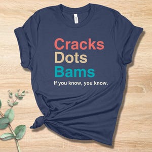 May include: A navy blue t-shirt with the text "Cracks Dots Bams" in colorful letters. The text is followed by the phrase "If you know, you know."