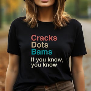 May include: Black t-shirt with the text "Cracks Dots Bams If you know, you know" in colorful letters.