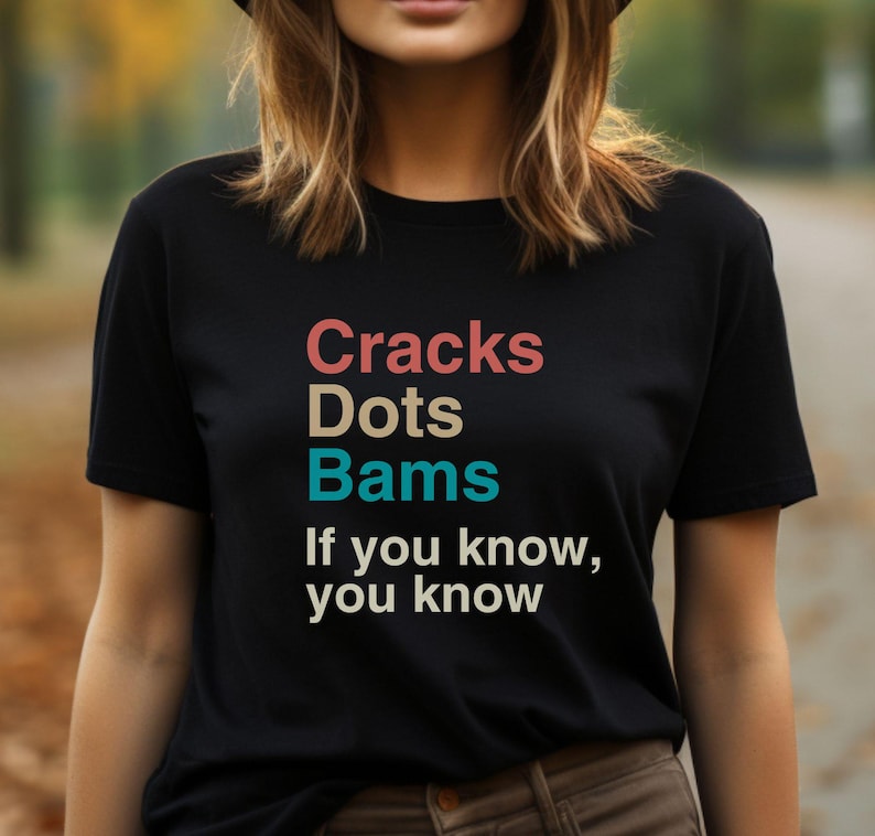 May include: Black t-shirt with the text "Cracks Dots Bams If you know, you know" in colorful letters.