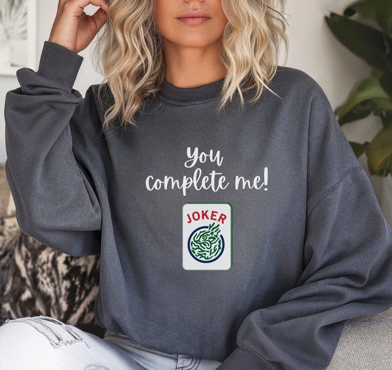 Puede incluir: Una sudadera gris con el texto "You complete me!" y un gr&aacute;fico de una carta de juego con la palabra "JOKER" en ella.