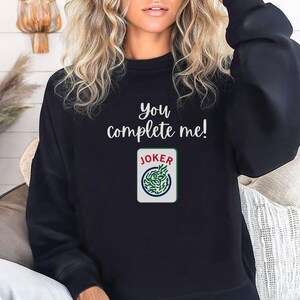Puede incluir: Sudadera negra con el texto "you complete me!" y un gr&aacute;fico de carta de joker verde y blanco.