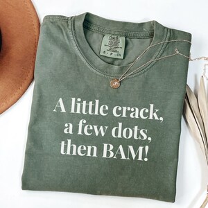 May include: An olive green t-shirt with the text "A little crack, a few dots, then BAM!" in white. A gold necklace with a pendant is on the shirt. The shirt is folded on a white surface.