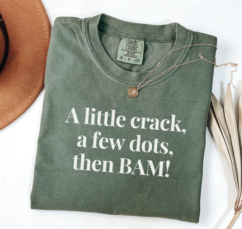 May include: An olive green t-shirt with the text "A little crack, a few dots, then BAM!" in white. A gold necklace with a pendant is on the shirt. The shirt is folded on a white surface.