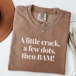 May include: Brown Comfort Colors t-shirt with the text "A little crack, a few dots, then BAM!" in white. A silver necklace with a small pendant is draped across the shirt. The shirt is size S-P-CH.