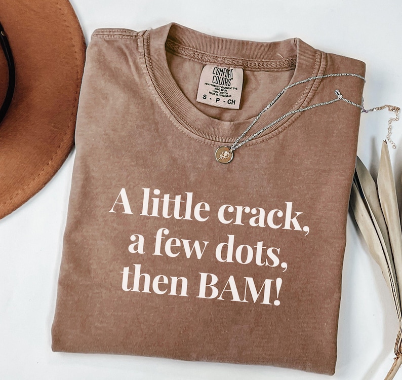 May include: Brown Comfort Colors t-shirt with the text "A little crack, a few dots, then BAM!" in white. A silver necklace with a small pendant is draped across the shirt. The shirt is size S-P-CH.