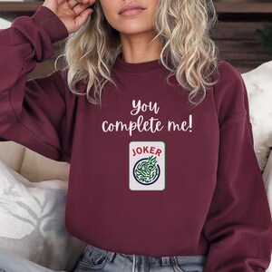Puede incluir: Una sudadera color burdeos con el texto "You complete me!" y un gr&aacute;fico de una carta de juego con la palabra "JOKER" en ella.