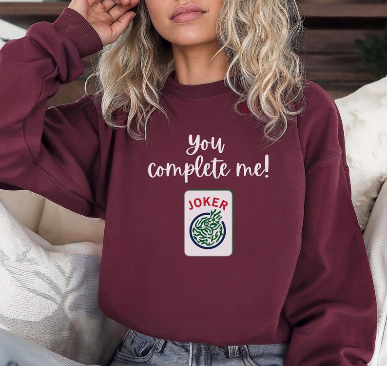 Puede incluir: Una sudadera color burdeos con el texto "You complete me!" y un gr&aacute;fico de una carta de juego con la palabra "JOKER" en ella.