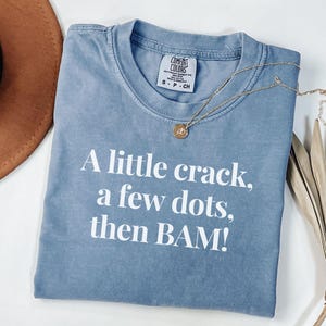 May include: A blue t-shirt with the text "A little crack, a few dots, then BAM!" in white. The shirt has a crew neck and a small tag near the collar. A gold necklace with a pendant is draped across the shirt.