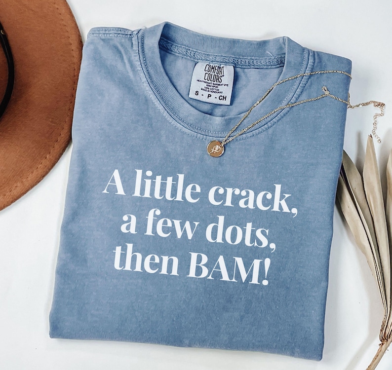 May include: A blue t-shirt with the text "A little crack, a few dots, then BAM!" in white. The shirt has a crew neck and a small tag near the collar. A gold necklace with a pendant is draped across the shirt.