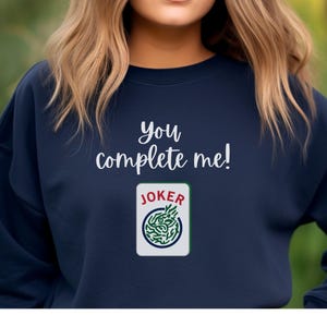 Puede incluir: Una sudadera azul marino con el texto "You complete me!" y un gr&aacute;fico de una carta de joker con un dise&ntilde;o verde y blanco.