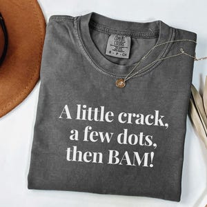 May include: Dark grey Comfort Colors t-shirt with the text "A little crack, a few dots, then BAM!" in white. A gold necklace with a pendant is on the shirt. A brown hat is partially visible in the upper left corner.