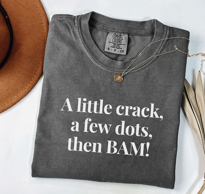May include: Dark grey Comfort Colors t-shirt with the text "A little crack, a few dots, then BAM!" in white. A gold necklace with a pendant is on the shirt. A brown hat is partially visible in the upper left corner.
