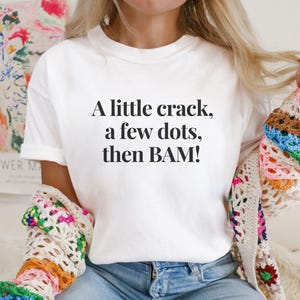 May include: White t-shirt with the text "A little crack, a few dots, then BAM!" in black. The shirt is paired with blue jeans and a colorful crocheted shawl. The overall aesthetic is casual and comfortable.