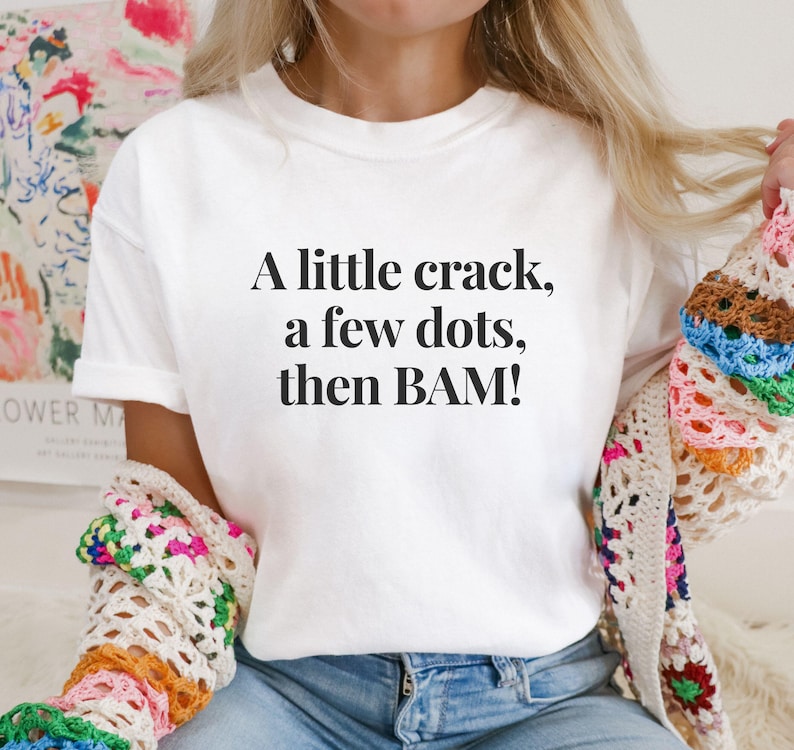 May include: White t-shirt with the text "A little crack, a few dots, then BAM!" in black. The shirt is paired with blue jeans and a colorful crocheted shawl. The overall aesthetic is casual and comfortable.