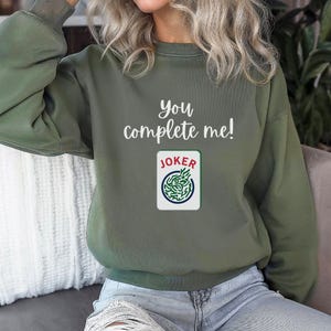Puede incluir: Una sudadera verde con el texto "you complete me!" y un gr&aacute;fico de carta de joker.