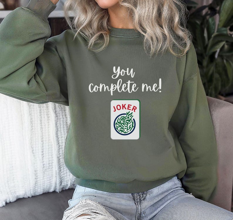 Puede incluir: Una sudadera verde con el texto "you complete me!" y un gr&aacute;fico de carta de joker.