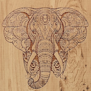 May include: A detailed wood carving of an elephant's head, featuring intricate patterns and designs. The elephant's face is the focal point, with large ears and a detailed trunk. The carving is set against a light wood grain background.