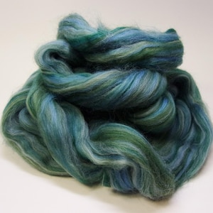 May include: A pile of teal, blue, and green fiber. The fiber is soft and fluffy, with a mix of colors creating a marbled effect. The fibers are tightly packed together, forming a dense mass.