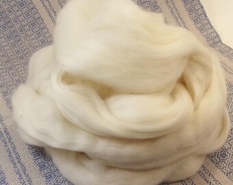 May include: A large, fluffy mass of undyed, off-white wool roving. The wool is soft and appears to be ready for spinning or felting. The background is a light blue and white patterned fabric.