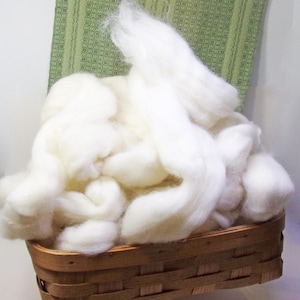 May include: A woven basket filled with fluffy, off-white wool fibers. The basket is made of light brown wood and has a rectangular shape. The wool appears soft and is piled high, suggesting a craft supply.