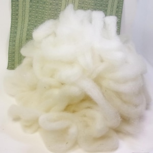 May include: A large pile of fluffy, off-white wool fibers. The wool has a soft, cloud-like appearance. A green and white patterned fabric is visible in the background. This image is suitable for craft projects.