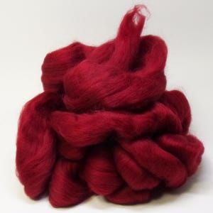 May include: A pile of vibrant red wool roving, tightly packed and textured. The fibers are soft and fluffy, ideal for various fiber art projects like needle felting or arm knitting. The deep red color is consistent throughout the wool.