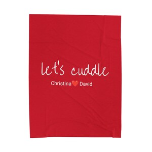 Puede incluir: Una manta roja con las palabras "let's cuddle" en escritura blanca. Debajo del texto están los nombres "Christina" y "David" separados por un símbolo de corazón rojo. La manta es de color rojo liso.