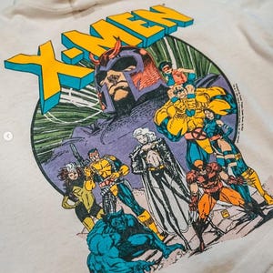 Peut inclure: T-shirt X-Men de couleur crème avec un graphisme vibrant des personnages X-Men, dont Magneto, Beast et Storm. Le logo X-MEN est jaune et bleu. Le design est de style bande dessinée vintage.