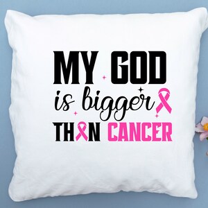 Puede incluir: Coj&iacute;n cuadrado blanco con el texto "MY GOD is bigger THAN CANCER" en negro y rosa. Un lazo rosa est&aacute; incluido en el dise&ntilde;o. El coj&iacute;n est&aacute; sobre una superficie azul claro.