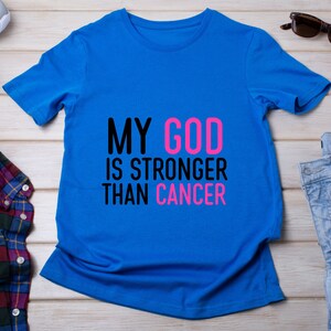 Puede incluir: Una camiseta azul brillante con el texto "MY GOD IS STRONGER THAN CANCER" en negro y rosa. La camiseta se muestra sobre una superficie de madera clara, con gafas de sol y vaqueros en el fondo.