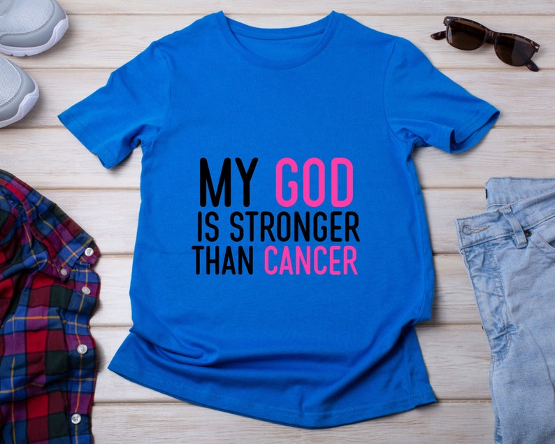 Puede incluir: Una camiseta azul brillante con el texto "MY GOD IS STRONGER THAN CANCER" en negro y rosa. La camiseta se muestra sobre una superficie de madera clara, con gafas de sol y vaqueros en el fondo.