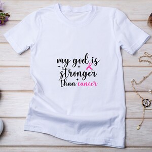 Puede incluir: Camiseta blanca con el texto "my god is stronger than cancer" en negro y rosa. El dise&ntilde;o incluye un lazo rosa, s&iacute;mbolo de la concienciaci&oacute;n sobre el c&aacute;ncer de mama. La camiseta se muestra sobre una superficie de madera clara.