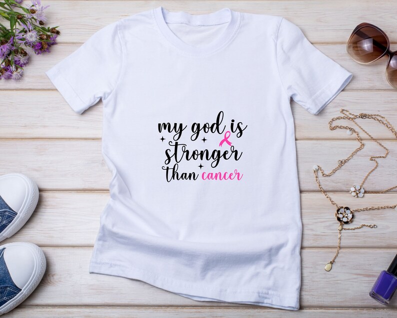 Puede incluir: Camiseta blanca con el texto "my god is stronger than cancer" en negro y rosa. El dise&ntilde;o incluye un lazo rosa, s&iacute;mbolo de la concienciaci&oacute;n sobre el c&aacute;ncer de mama. La camiseta se muestra sobre una superficie de madera clara.