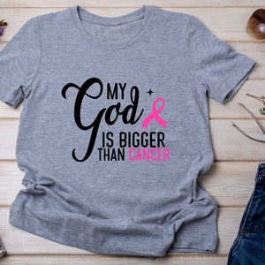 Puede incluir: Camiseta gris jaspeado con el texto "My God is Bigger Than Cancer" en negro y rosa. Tambi&eacute;n incluye un lazo rosa. La camiseta es una prenda de vestir informal.