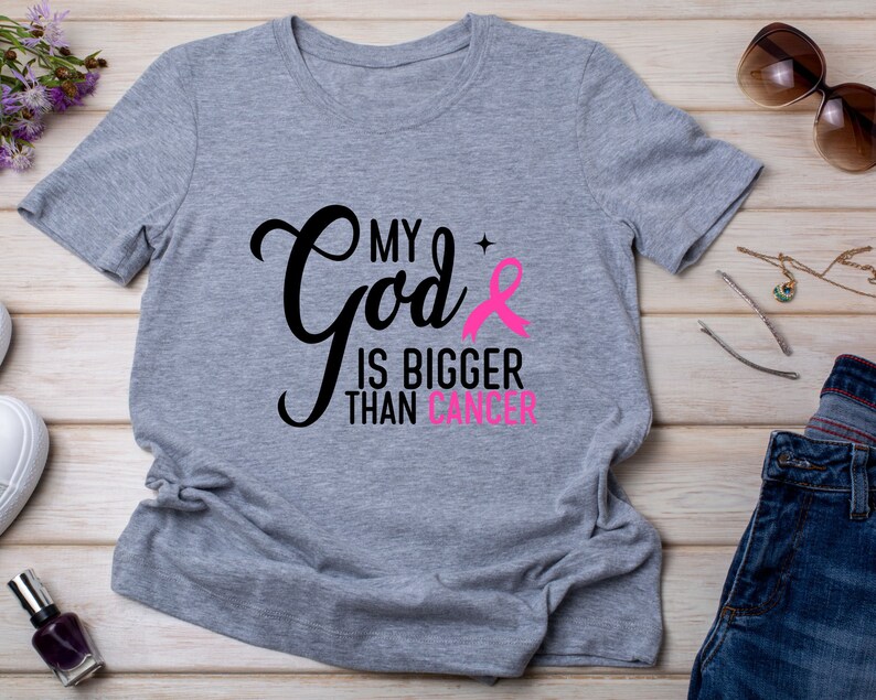 Puede incluir: Camiseta gris jaspeado con el texto "My God is Bigger Than Cancer" en negro y rosa. Tambi&eacute;n incluye un lazo rosa. La camiseta es una prenda de vestir informal.