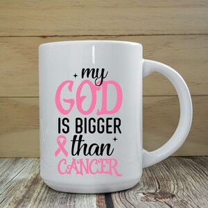 Puede incluir: Taza de cer&aacute;mica blanca con el texto "my GOD is BIGGER than CANCER" en rosa y negro. La taza presenta un s&iacute;mbolo de cinta rosa. La taza tiene un asa grande y est&aacute; sobre un fondo de madera.