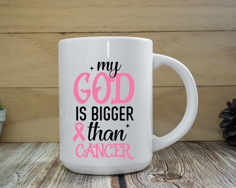 Puede incluir: Taza de cer&aacute;mica blanca con el texto "my GOD is BIGGER than CANCER" en rosa y negro. La taza presenta un s&iacute;mbolo de cinta rosa. La taza tiene un asa grande y est&aacute; sobre un fondo de madera.