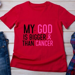Puede incluir: Una camiseta roja con el texto "MY GOD IS BIGGER THAN CANCER" en negro y rosa. Se incluye un lazo rosa. La camiseta se muestra sobre una superficie de madera clara con vaqueros azules, una camisa a cuadros y zapatillas.