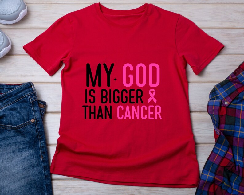 Puede incluir: Una camiseta roja con el texto "MY GOD IS BIGGER THAN CANCER" en negro y rosa. Se incluye un lazo rosa. La camiseta se muestra sobre una superficie de madera clara con vaqueros azules, una camisa a cuadros y zapatillas.