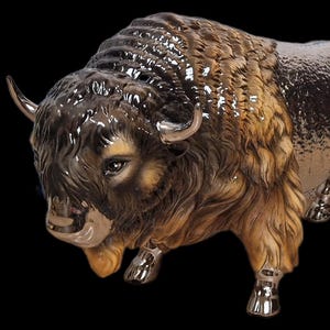 May include: A ceramic bison figurine with a detailed design. The sculpture features a mix of brown, tan, and silver tones, with a textured fur effect. The bison has curved horns and is posed in a standing position, showcasing its powerful build.