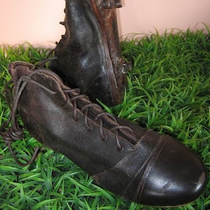 Può includere: Due scarpini da calcio vintage in pelle con lacci. Gli scarpini sono marroni e hanno un aspetto usurato. Sono su un letto di erba verde.