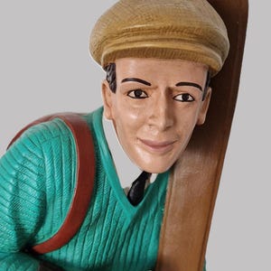 May include: A vintage figurine of a skier, wearing a green sweater, brown gloves, and a tan cap. The skier is holding a pair of wooden skis and a walking stick. The figurine has a red backpack and a black tie.