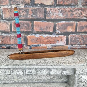 Könnte beinhalten: Ein Holzwebstuhl mit einem blau-rot gestreiften Faden, der um den Schussfaden gewickelt ist. Der Webstuhl steht auf einer weißen, abgenutzten Oberfläche.