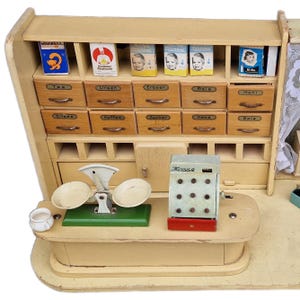 May include: A vintage wooden toy shop with a counter, drawers labeled with German food items, and a cash register. The shop has a scale, small boxes, and a shelf with various products. The overall color scheme is cream and light brown.