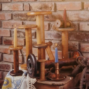 Op de afbeelding: Een verzameling houten spoelen die zijn omgevormd tot kaarsenhouders. De spoelen zijn gerangschikt op een houten oppervlak met een bakstenen muur op de achtergrond. De kaarsen zijn aangestoken en de vlammen zijn zichtbaar.