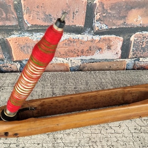 Könnte beinhalten: Ein Holzschiffchen mit einem roten und weißen Garnfaden, der darin eingesetzt ist. Das Schiffchen wird zum Weben verwendet.