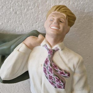 Könnte beinhalten: Eine Keramikfigur eines Mannes, der ein weißes Hemd mit einer pinkfarbenen und grünen Blümchen-Krawatte trägt. Er lächelt und hat seinen Arm erhoben, als ob er etwas halten würde.