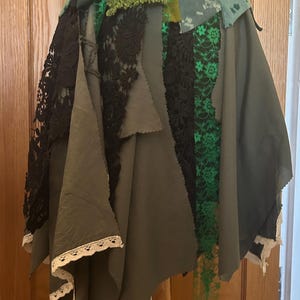 May include: A layered skirt featuring olive green fabric panels, black floral lace overlays, and bright green lace accents. The waistband is adorned with a wide white lace trim. The hemline is uneven and finished with a delicate cream-colored lace border.