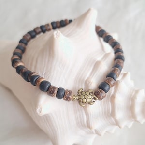 Pode incluir: Pulseira de contas de madeira castanha escura e preta alternadas, com um pequeno berloque de tartaruga dourada. A pulseira é exibida em uma concha branca. As contas têm aproximadamente 0,5 cm de diâmetro.